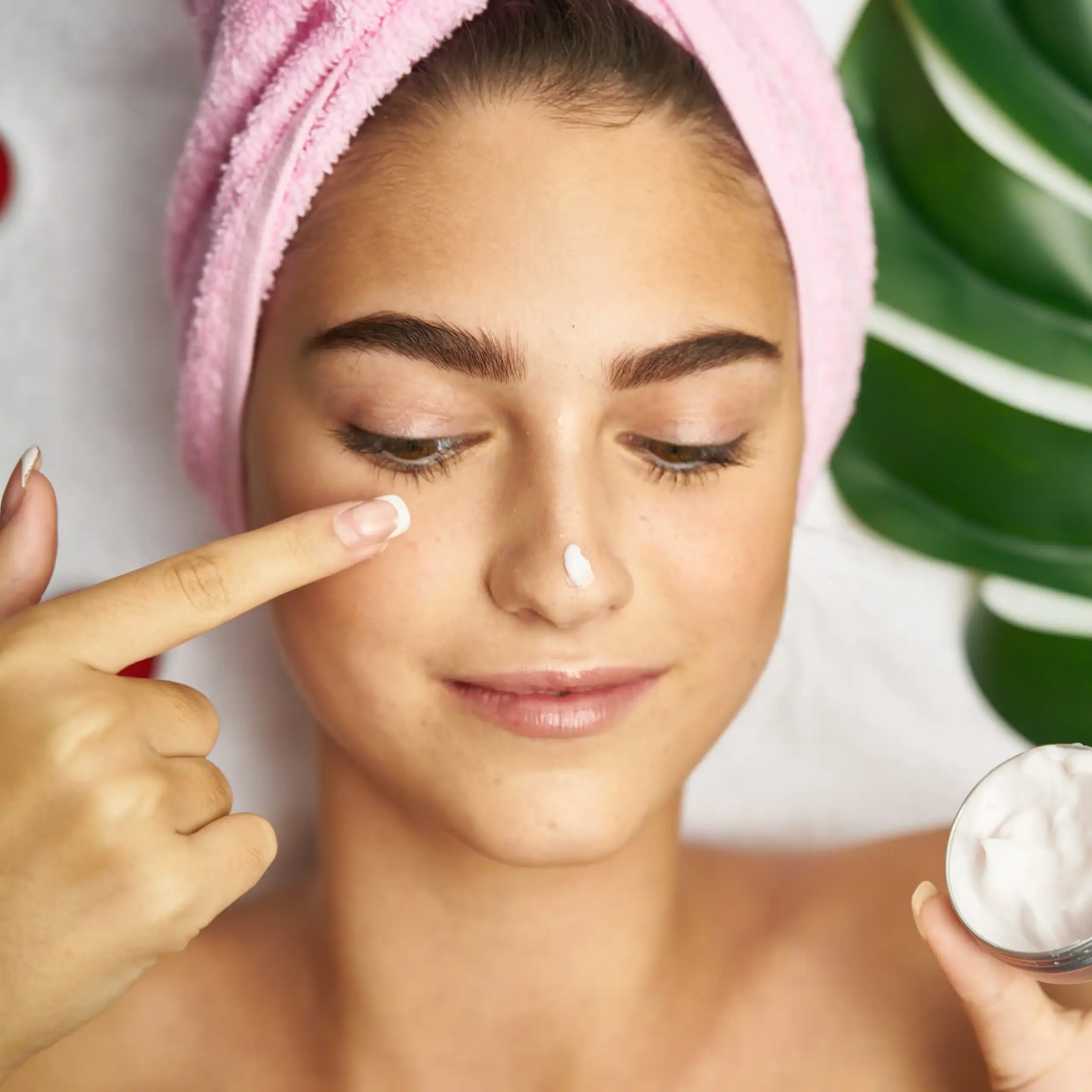 Woman with hair wrapped in pink towel applies a topical treatment to her face